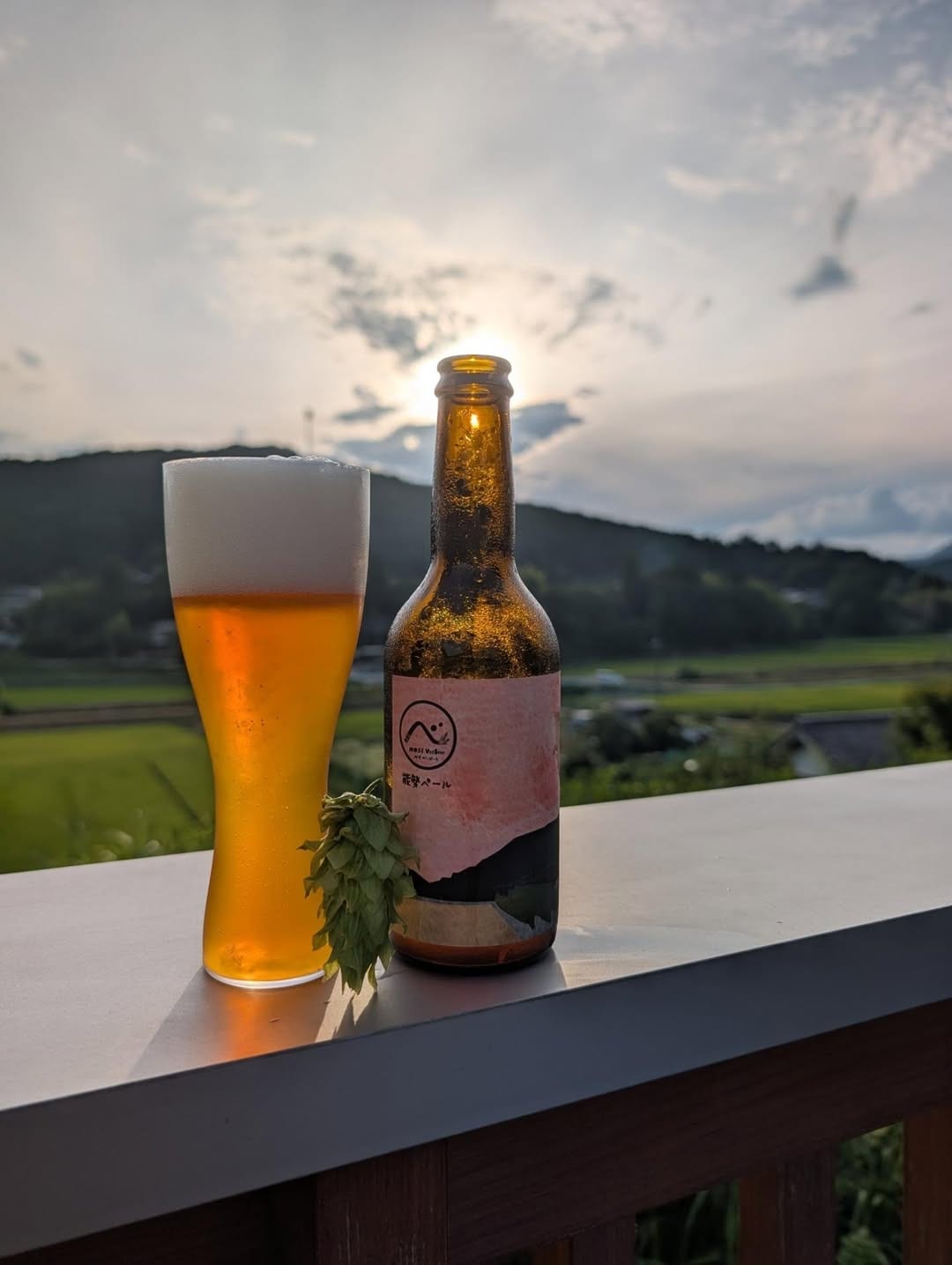 夕暮れの田んぼを背に、手すりの上に置かれた瓶と、注いだクラフトビールのグラス。足元にホップの毬花。